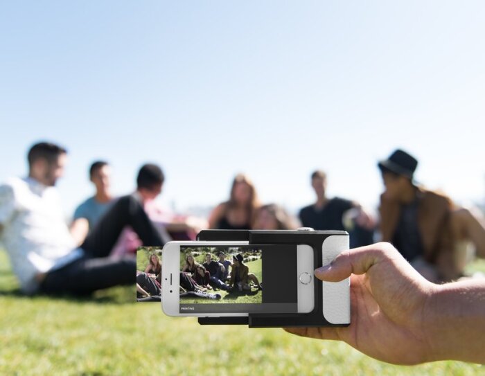 Une main tient une imprimante photo Prynt sur un smartphone, qui est en train d'imprimer la photo d'un groupe de jeunes gens assis dans l'herbe en extérieur