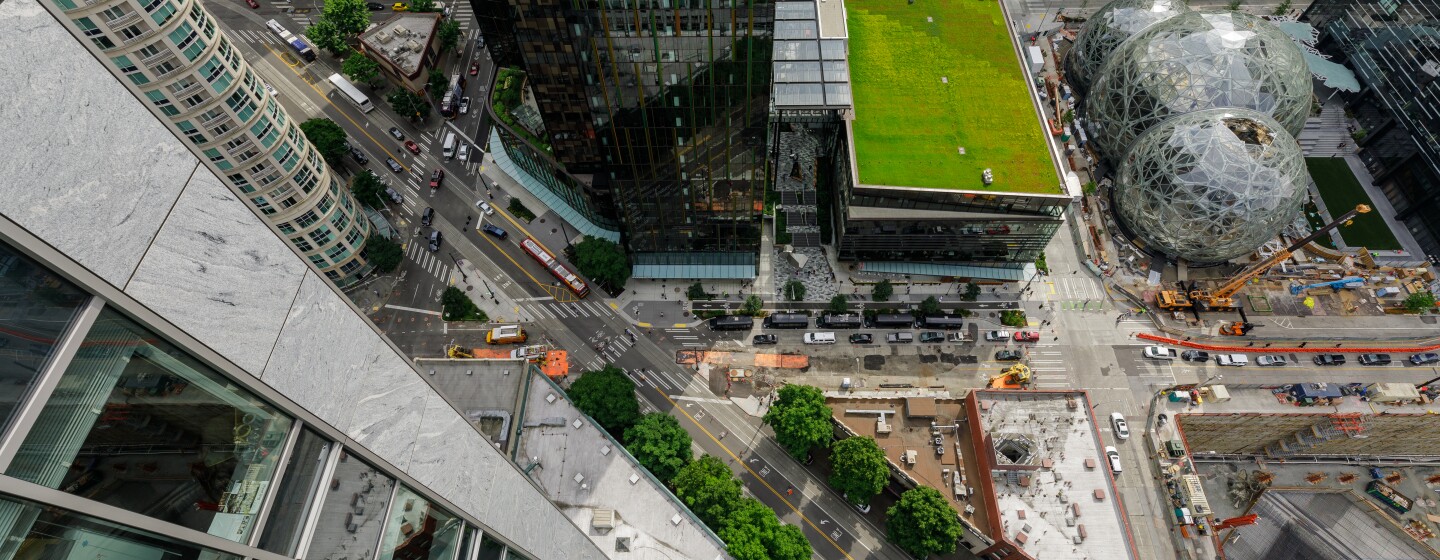 A view from on top of a skyscraper, looking down on the development of the Amazon campus. A crane operator directs machinery near the Amazon Spheres. Cars drive on urban streets.