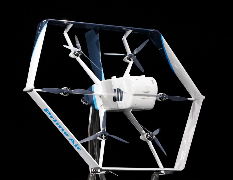 A white and light blue Prime Air drone in front of a black background. Six propellers and a main body are visible in the center of the drone.