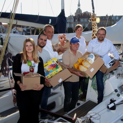 Eine Gruppe Menschen steht auf einem kleinen Segelboot. JEder hält einen Karton mit Spielzeug und Kuscheltieren in der Hand.