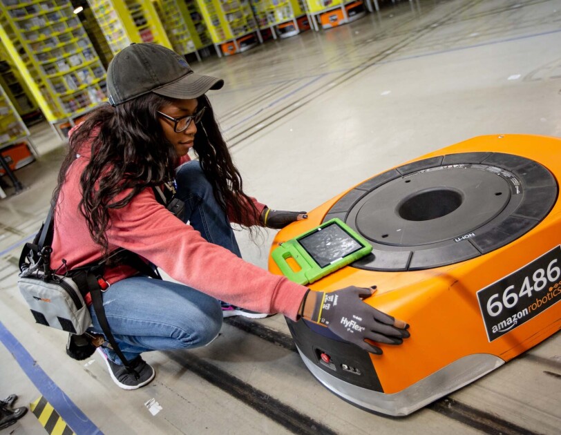 A woman kneels next to a robot, while also working on a tablet, at a Columbus, Ohio fulfillment center, CMH1