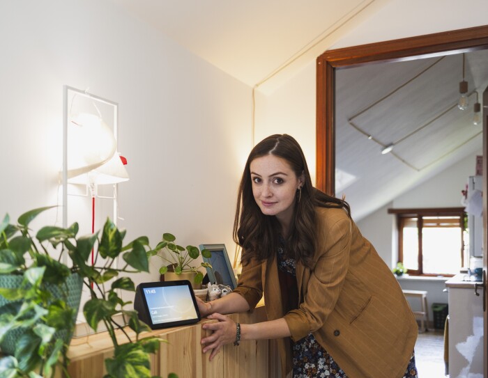 Elisa Reale, al centro della foto, è chinata verso un mobile. Guarda in camera, vicino alla mano destra ha un dispositivo Echo Show con lo schermo acceso. In primo piano, posata sullo stesso mobile, si vede una pianta in un vaso verde. Elisa indossa una giacca beige a righe bianche, ha i capelli lunghi e mossi. Sotto la giacca indossa una gonna a fiori colorata.